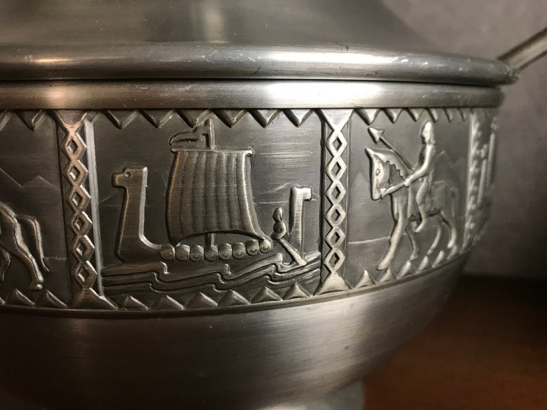 Puede incluir: Un taz&oacute;n de servir de peltre plateado con tapa, que presenta un dise&ntilde;o en relieve de un barco vikingo, un jinete y un patr&oacute;n geom&eacute;trico.