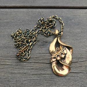 May include: A gold-toned necklace with a large, abstract pendant. The pendant is made of two overlapping leaves with a textured surface and a series of small circles.