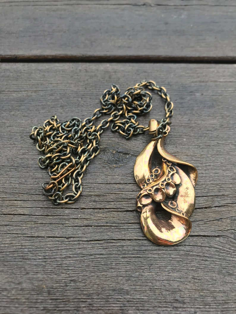 May include: A gold-toned necklace with a large, abstract pendant. The pendant is made of two overlapping leaves with a textured surface and a series of small circles.