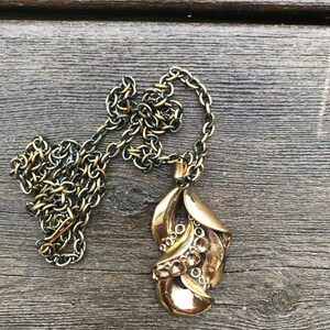 May include: A gold-toned necklace with a detailed pendant shaped like a stylized leaf. The pendant has a textured surface and a series of small circles.