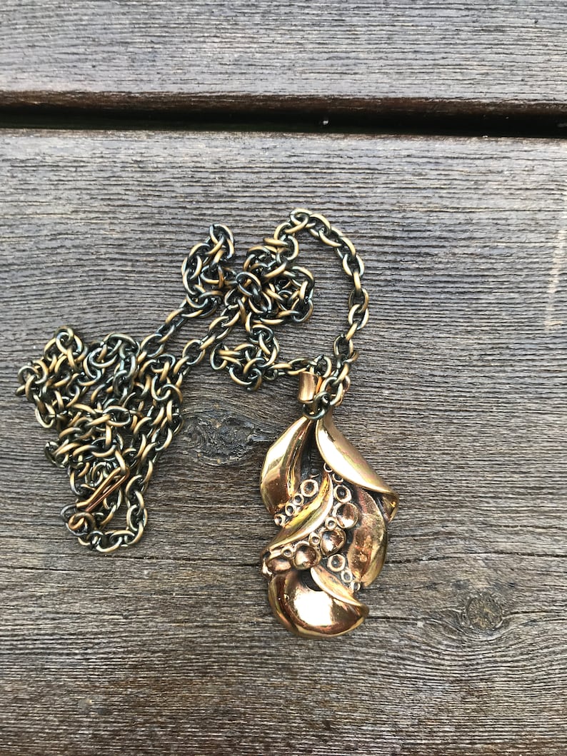 May include: A gold-toned necklace with a detailed pendant shaped like a stylized leaf. The pendant has a textured surface and a series of small circles.