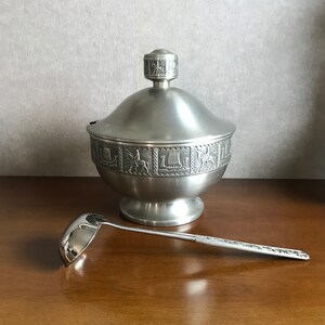 Puede incluir: Un azucarero de peltre plateado con tapa y cuchar&oacute;n a juego. El bol est&aacute; decorado con un intrincado dise&ntilde;o de figuras y animales.