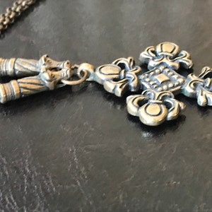 Puede incluir: Un collar dorado con un colgante de cruz adornado con detalles. La cruz tiene un centro cuadrado con un patr&oacute;n decorativo y cuatro dise&ntilde;os de flor de lis.