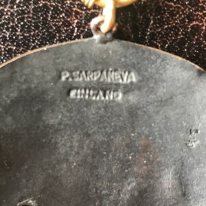 K&ouml;nnte beinhalten: Ein silberfarbener Metallanh&auml;nger mit einem erhabenen Design. Die R&uuml;ckseite des Anh&auml;ngers ist mit dem Text "P.SARDANEVA ENGLAND" graviert.