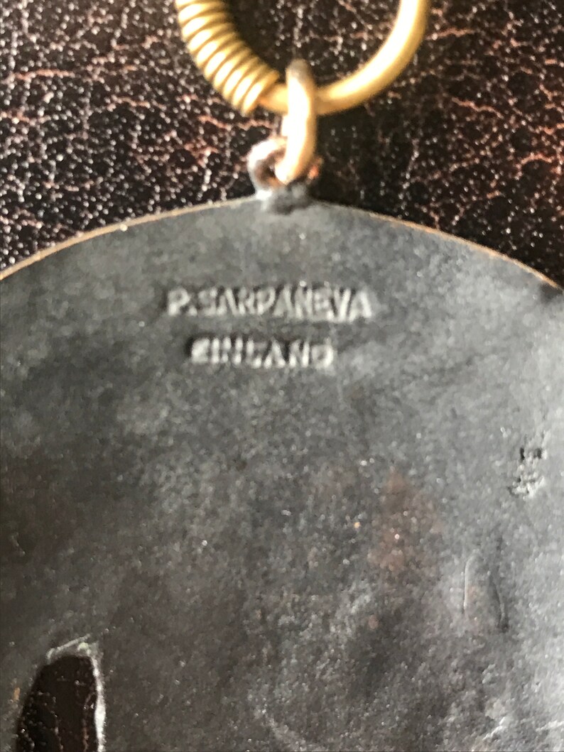 K&ouml;nnte beinhalten: Ein silberfarbener Metallanh&auml;nger mit einem erhabenen Design. Die R&uuml;ckseite des Anh&auml;ngers ist mit dem Text "P.SARDANEVA ENGLAND" graviert.