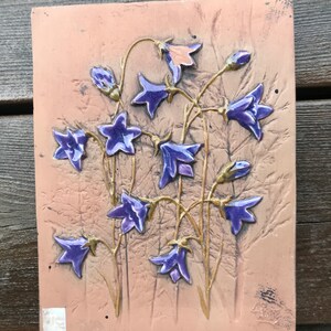 May include: A ceramic tile featuring a raised design of purple bellflowers on a light brown background.