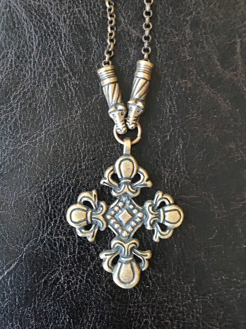 Puede incluir: Un collar dorado con un colgante de cruz detallado y dos extremos de cadena decorativos. La cruz tiene un centro en forma de diamante y dise&ntilde;os florales intrincados.