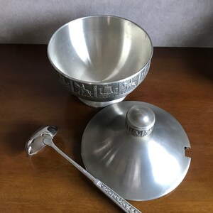 Puede incluir: Un bol de servir de plata con tapa y cuchar&oacute;n. El bol y la tapa tienen un dise&ntilde;o en relieve alrededor del borde. El cuchar&oacute;n tiene un mango decorativo.