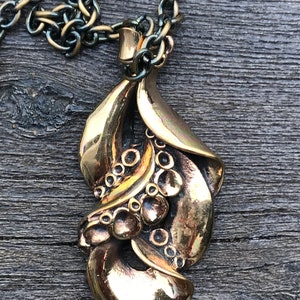 May include: A gold-toned pendant necklace with a detailed, ornate design featuring a series of rounded shapes and a leaf-like form. The pendant is attached to a black and gold chain.