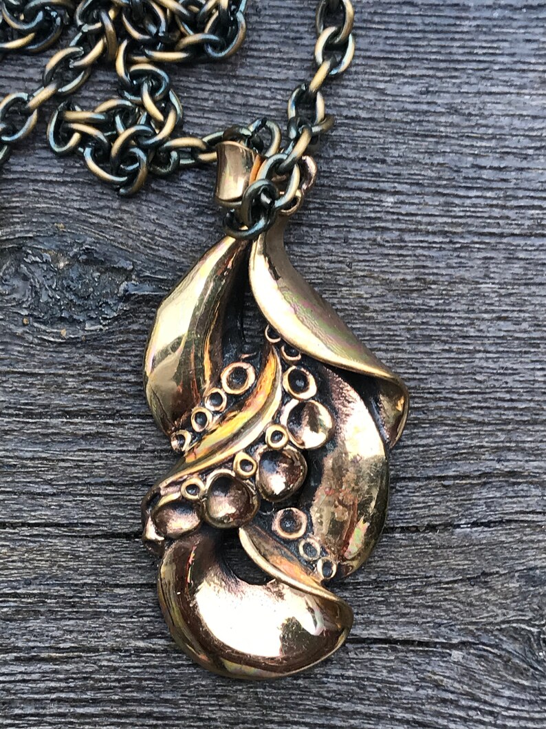 May include: A gold-toned pendant necklace with a detailed, ornate design featuring a series of rounded shapes and a leaf-like form. The pendant is attached to a black and gold chain.