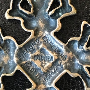 Puede incluir: Un colgante de cruz de metal plateado con detalles intrincados. La parte posterior del colgante est&aacute; grabada con el texto "GIGANT, ENGLAND, ORIGINAL, MADE IN NORWAY".