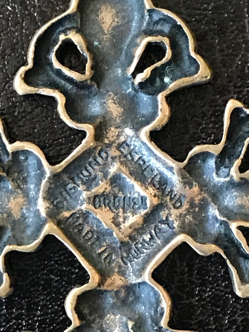 Puede incluir: Un colgante de cruz de metal plateado con detalles intrincados. La parte posterior del colgante est&aacute; grabada con el texto "GIGANT, ENGLAND, ORIGINAL, MADE IN NORWAY".