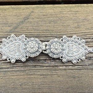 Può includere: Una chiusura color argento con un design ornato. La chiusura è composta da due metà simmetriche, ciascuna con un centro a forma di paisley e un motivo floreale. La chiusura è collegata da una piccola cerniera, su uno sfondo di legno.