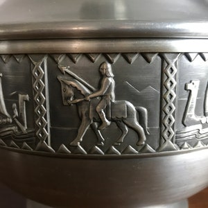 Puede incluir: Un taz&oacute;n de metal plateado con un dise&ntilde;o decorativo que presenta un caballero a caballo y dos barcos. El taz&oacute;n tiene una superficie lisa y brillante.