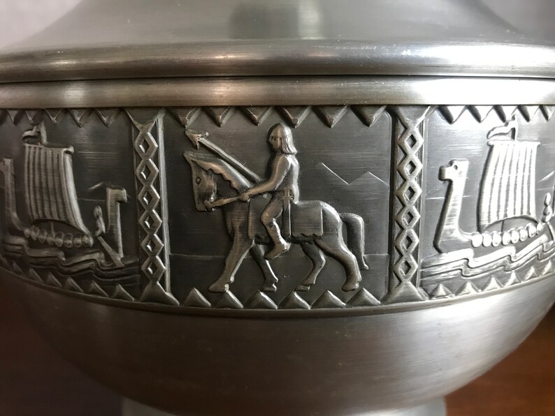 Puede incluir: Un taz&oacute;n de metal plateado con un dise&ntilde;o decorativo que presenta un caballero a caballo y dos barcos. El taz&oacute;n tiene una superficie lisa y brillante.