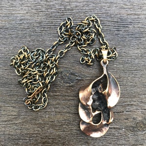 May include: A bronze pendant necklace with a unique abstract design. The pendant is made of multiple overlapping pieces of metal, creating a flowing, organic shape. The chain is a simple, silver-toned chain.