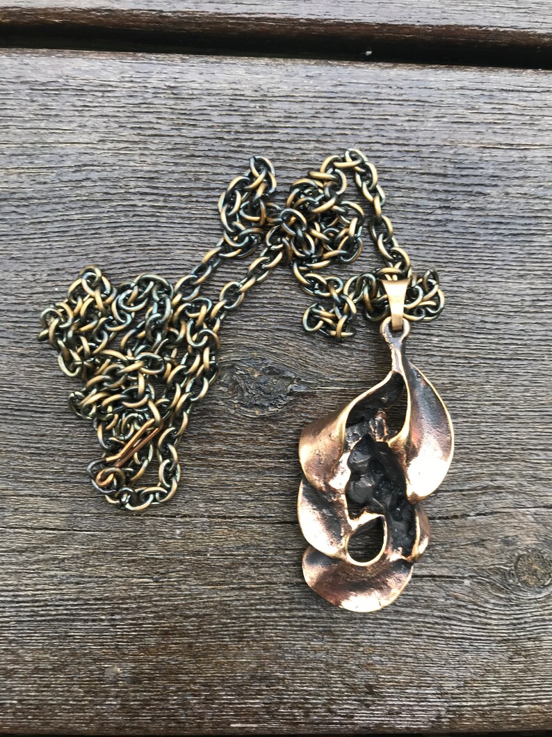 May include: A bronze pendant necklace with a unique abstract design. The pendant is made of multiple overlapping pieces of metal, creating a flowing, organic shape. The chain is a simple, silver-toned chain.