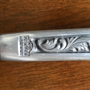 Puede incluir: Una cuchara decorativa de plata adornada con un dise&ntilde;o floral. La cuchara tiene las palabras "H.S. PEWTER NORWAY" grabadas en el mango.