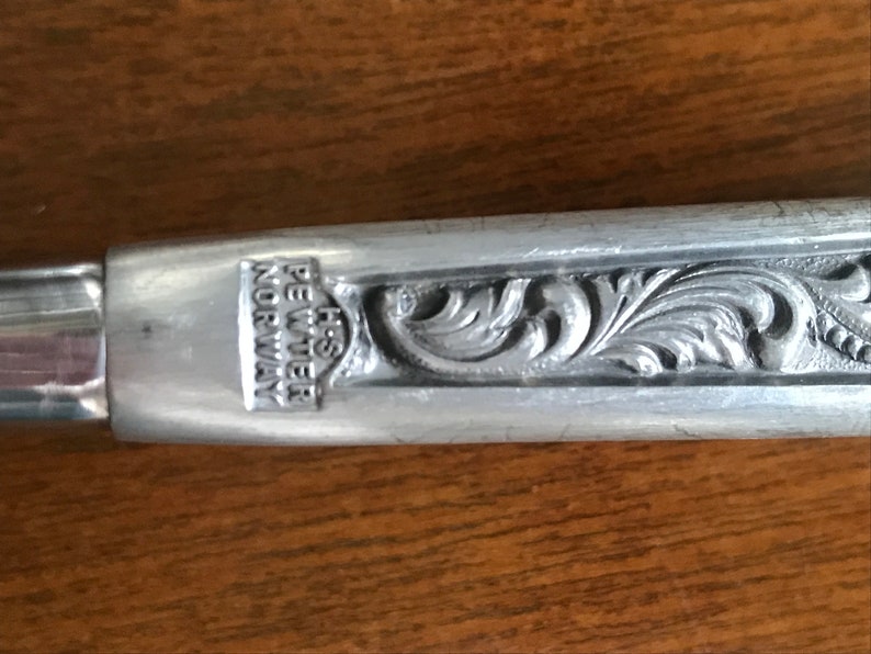 Puede incluir: Una cuchara decorativa de plata adornada con un dise&ntilde;o floral. La cuchara tiene las palabras "H.S. PEWTER NORWAY" grabadas en el mango.