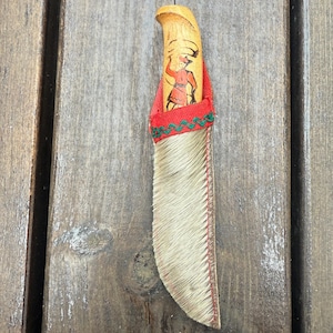 Pode incluir: Uma pequena faca com cabo de madeira com uma figura pintada em vermelho e castanho. A lâmina é coberta com pele castanha e costuras vermelhas. A faca está sobre uma superfície de madeira.