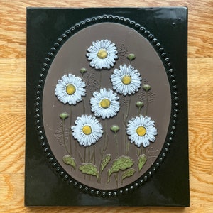 May include: A brown ceramic tile with a black frame and an oval-shaped design of white daisies with yellow centres. The tile has a raised, textured surface and is decorated with a small signature in the bottom right corner.