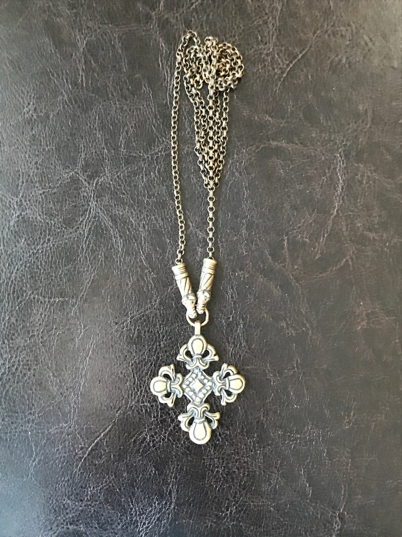 Puede incluir: Un collar de cadena de plata con un colgante de cruz de plata. La cruz tiene un dise&ntilde;o detallado con un peque&ntilde;o diamante en el centro.