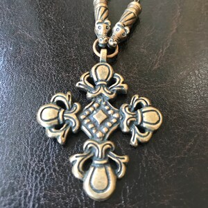 Puede incluir: Un collar dorado con un gran colgante de cruz adornado. La cruz tiene un centro en forma de diamante y detalles intrincados. El collar tiene dos cuentas decorativas en la parte superior.