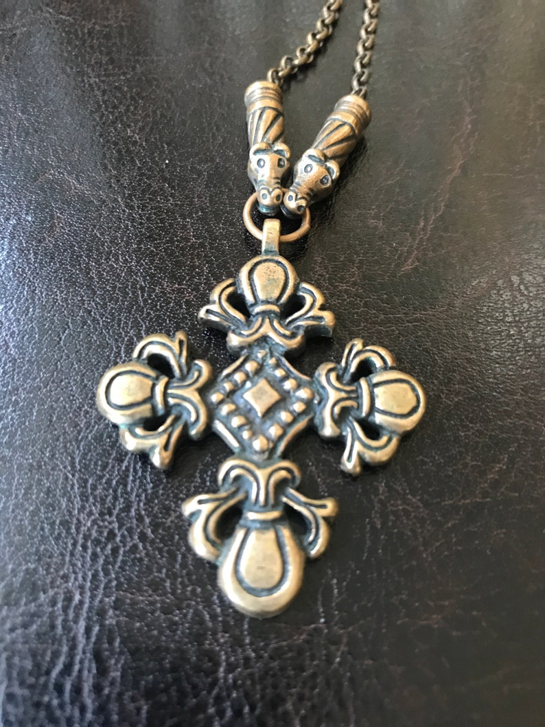 Puede incluir: Un collar dorado con un gran colgante de cruz adornado. La cruz tiene un centro en forma de diamante y detalles intrincados. El collar tiene dos cuentas decorativas en la parte superior.