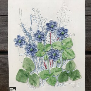 May include: A white ceramic tile featuring an embossed design of blue and green flowers. The tile is signed "Guno" in the bottom right corner and bears a "JIE Gantofta Sweden" logo in the bottom left corner.