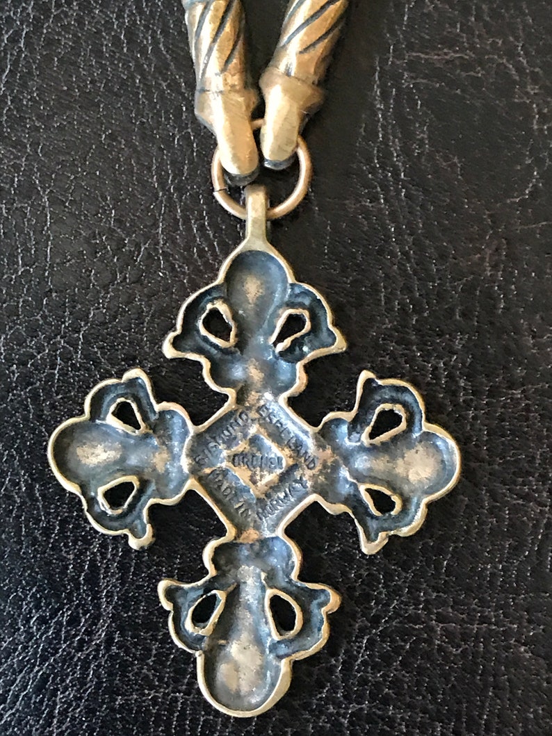 Puede incluir: Un colgante de cruz de metal dorado con detalles intrincados. La parte posterior del colgante tiene la inscripci&oacute;n "Gift from Espeland Original Made in Norway".