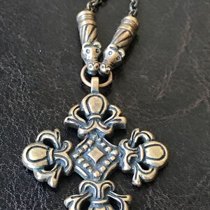 Puede incluir: Un collar dorado con un colgante de cruz. La cruz tiene un dise&ntilde;o detallado con un centro en forma de diamante y adornos ornamentados. El collar tiene dos cuentas decorativas en la parte superior.