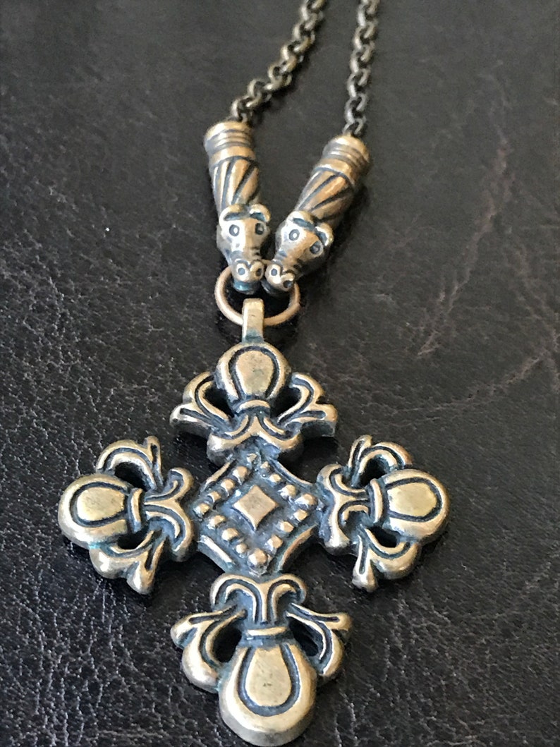 Puede incluir: Un collar dorado con un colgante de cruz. La cruz tiene un dise&ntilde;o detallado con un centro en forma de diamante y adornos ornamentados. El collar tiene dos cuentas decorativas en la parte superior.