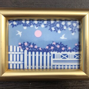 May include: A gold-framed picture with a blue and white ceramic tile depicting a white picket fence, two white birds, and pink flowers. The tile has a blue background with a white circle in the center.