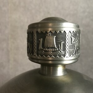 Puede incluir: Un tap&oacute;n de botella de peltre plateado con un dise&ntilde;o detallado de un barco vikingo y otras figuras. El tap&oacute;n es redondo y tiene una superficie lisa y pulida.