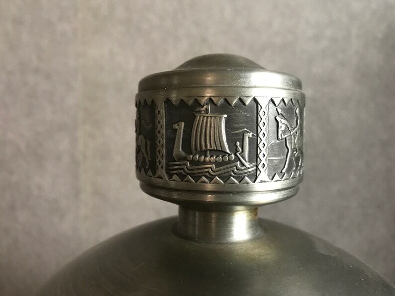 Puede incluir: Un tap&oacute;n de botella de peltre plateado con un dise&ntilde;o detallado de un barco vikingo y otras figuras. El tap&oacute;n es redondo y tiene una superficie lisa y pulida.