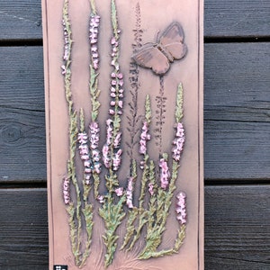May include: A brown ceramic wall plaque with a raised design of pink flowers and a brown butterfly. The plaque has a brown background and is signed "jic" in the bottom left corner.
