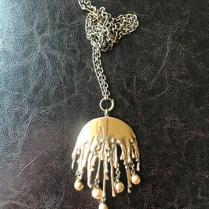 K&ouml;nnte beinhalten: Eine goldene Halskette mit einem gro&szlig;en, abstrakten Anh&auml;nger mit einem strukturierten, tropfenden Design und f&uuml;nf baumelnden runden Perlen.
