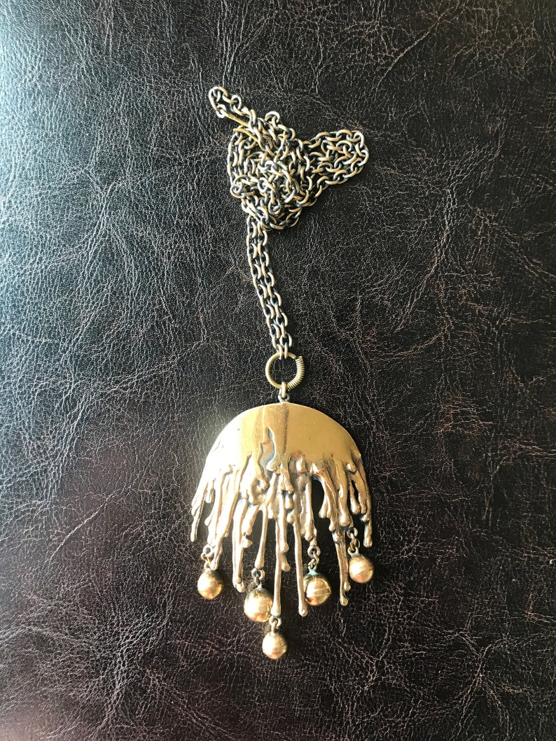 K&ouml;nnte beinhalten: Eine goldene Halskette mit einem gro&szlig;en, abstrakten Anh&auml;nger mit einem strukturierten, tropfenden Design und f&uuml;nf baumelnden runden Perlen.