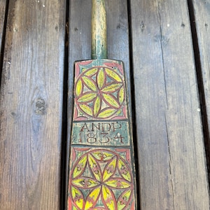 Könnte beinhalten: Ein antikes Butterfass aus Holz mit langem Griff und rechteckigem Paddelende. Das Paddelende zeigt ein Blumenmuster in Gelb, Rot und Grün mit der Inschrift "ANDP 1834". Der Griff ist grün und rot bemalt.