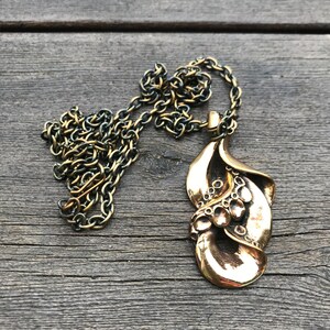 May include: A gold-toned necklace with a large, abstract pendant. The pendant is made of metal and has a textured, organic shape. The chain is a simple, linked chain.
