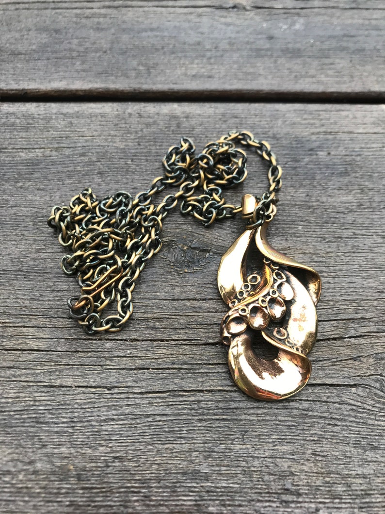 May include: A gold-toned necklace with a large, abstract pendant. The pendant is made of metal and has a textured, organic shape. The chain is a simple, linked chain.