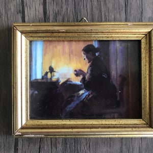 Könnte beinhalten: Ein gerahmtes Gemälde einer Frau, die in einem schwach beleuchteten Raum sitzt und bei Kerzenlicht näht. Das Gemälde ist in einem goldenen Rahmen gefasst.