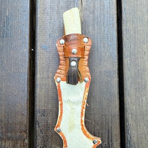 Peut inclure: Un étui de couteau en cuir fait à la main avec un couteau à manche en os. L'étui est en cuir bronzé avec une bordure orange et des rivets argentés. L'étui a une matière semblable à de la fourrure blanche et beige sur le devant.