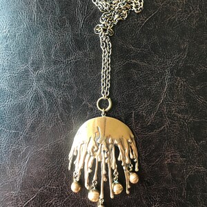 K&ouml;nnte beinhalten: Eine goldfarbene Halskette mit einem gro&szlig;en, abstrakten Anh&auml;nger in Tropfenform und mehreren runden, baumelnden Charms.