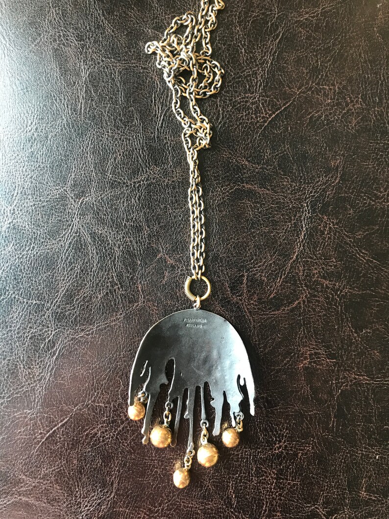 K&ouml;nnte beinhalten: Eine Halskette mit einem schwarzen Metall-Anh&auml;nger in Tropfenform und einer goldfarbenen Kette und Perlen. Der Anh&auml;nger tr&auml;gt auf der R&uuml;ckseite den Text "POMPADOUR MILANO".