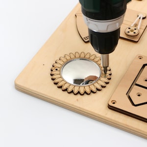 May include: A wooden activity board with a sunflower-shaped mirror being assembled with a power drill. The board features other wooden components, including a pulley and a geometric shape. The wood has a natural, light colour.