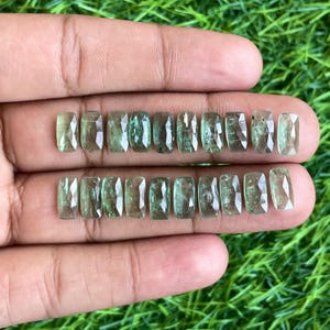 May include: A collection of 15 green gemstone rectangular cabochons, displayed on a hand. The gemstones are cut and polished, with a smooth, glassy surface.