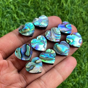May include: A collection of heart-shaped abalone shell cabochons. The iridescent shells display a range of blues, greens, and purples, with a black backing. The hearts are held in a hand against a green background.