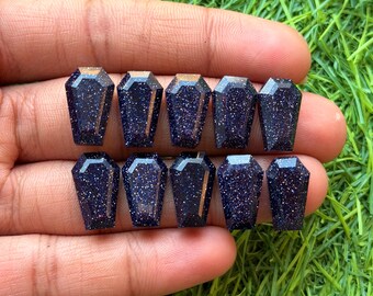 May include: Ten dark blue gemstone coffin-shaped beads with sparkling inclusions. The beads are held in a hand against a background of green grass. Each bead has a faceted top and a polished finish.