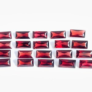 May include: A collection of twenty-one rectangular, deep red gemstones, possibly garnets, arranged on a white surface. Each gem has a polished, reflective surface and a rectangular shape, with beveled edges.